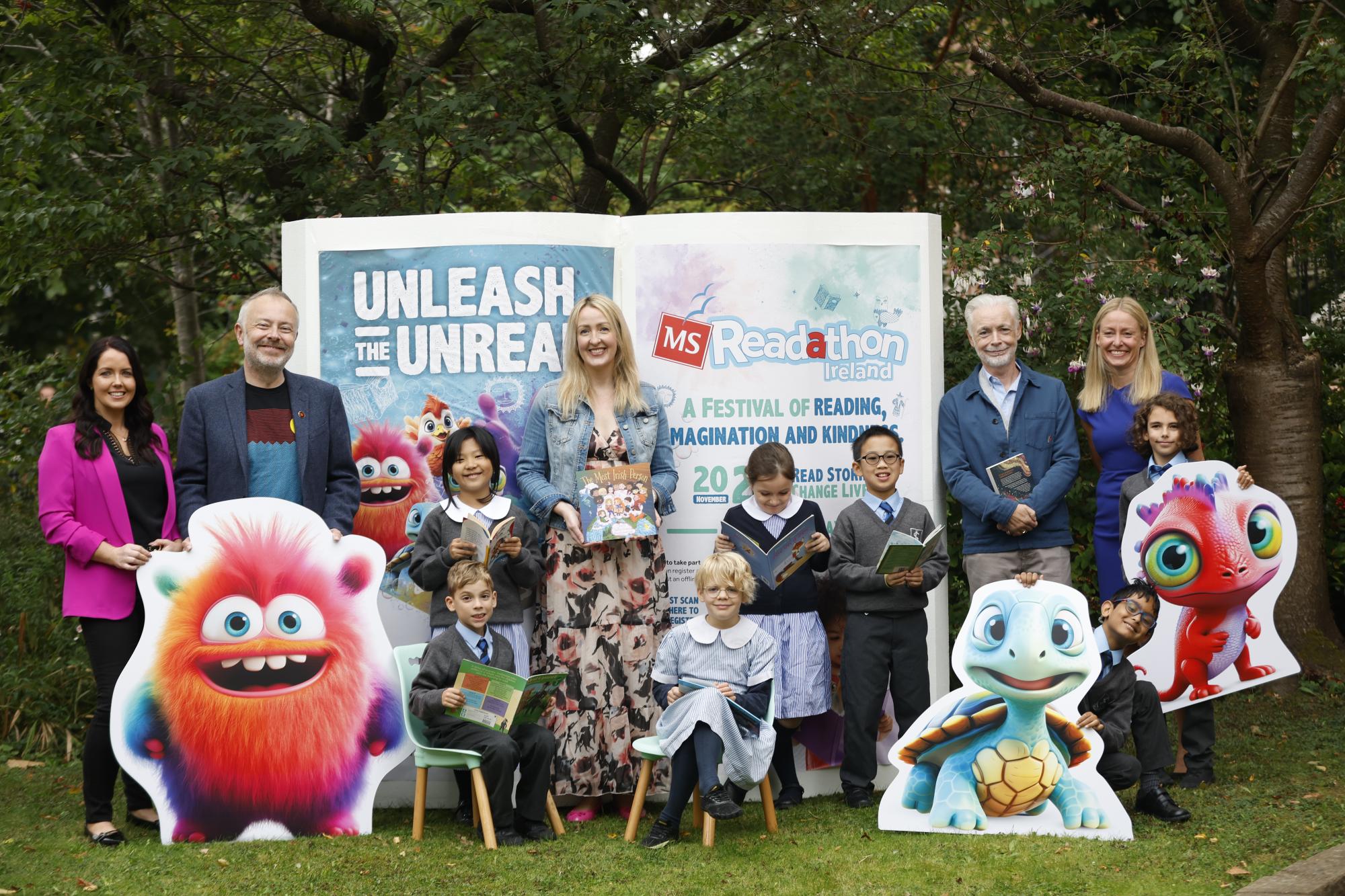 A giant, oversized book is open in the background, with a group of adults and children, participants in the MS Readathon launch, standing and sitting in front of it. Cardboard cutouts of various characters are placed around the group, adding a playful and literary theme to the scene. The group appears engaged, celebrating the kickoff of the reading initiative. 