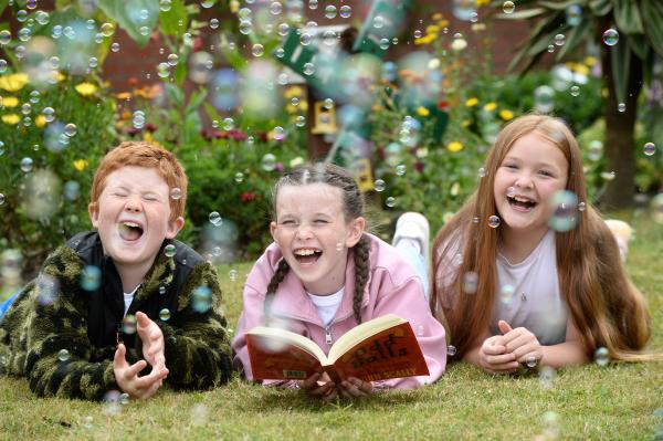 Three happy kids lying on the grass. One of them is holding and reading a book, while bubbles float around them