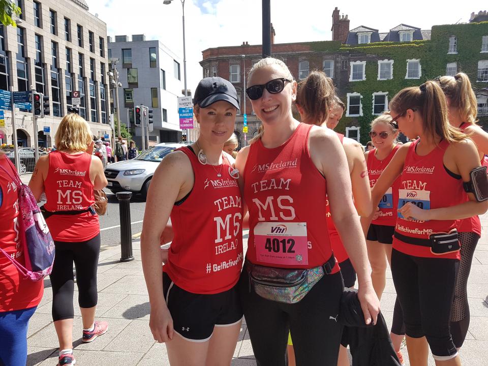 Two people smiling at the camera, with others in the background, all wearing red t-shirts in preparation for a mini marathon