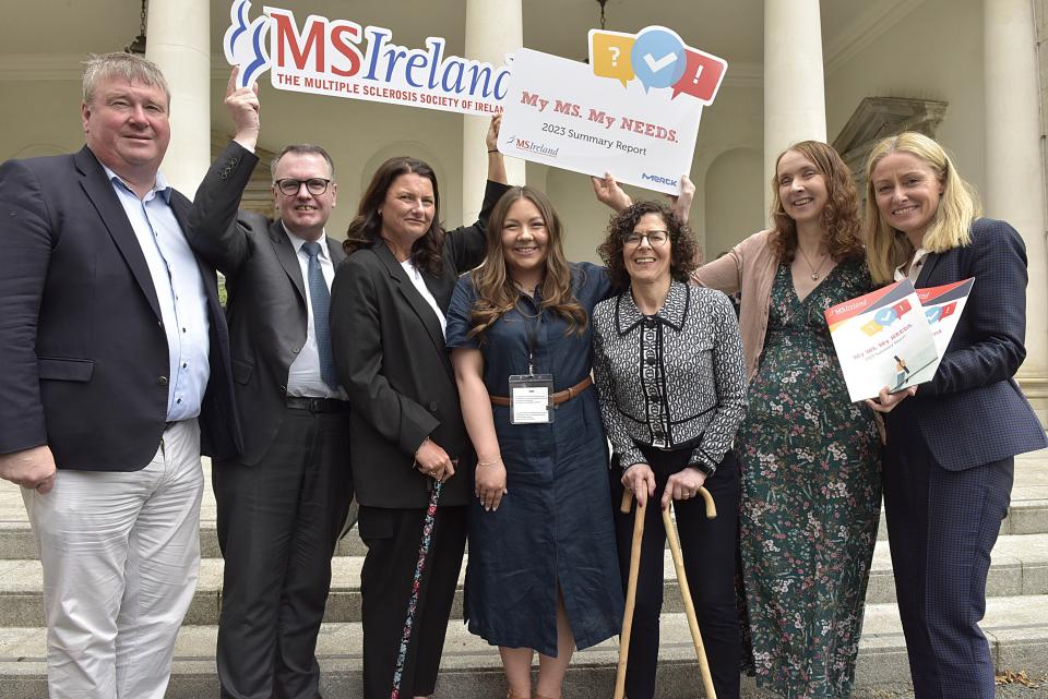 Seven individuals standing in a row, each holding banners and copies of survey results