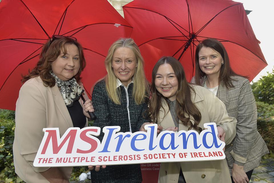 Four people standing together. Two of them are holding red umbrellas, while another holds a sign.