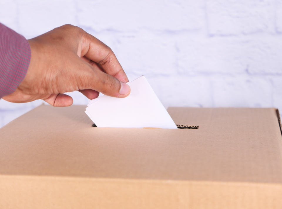 Hand placing a paper into a ballot box in front of a white brick wall