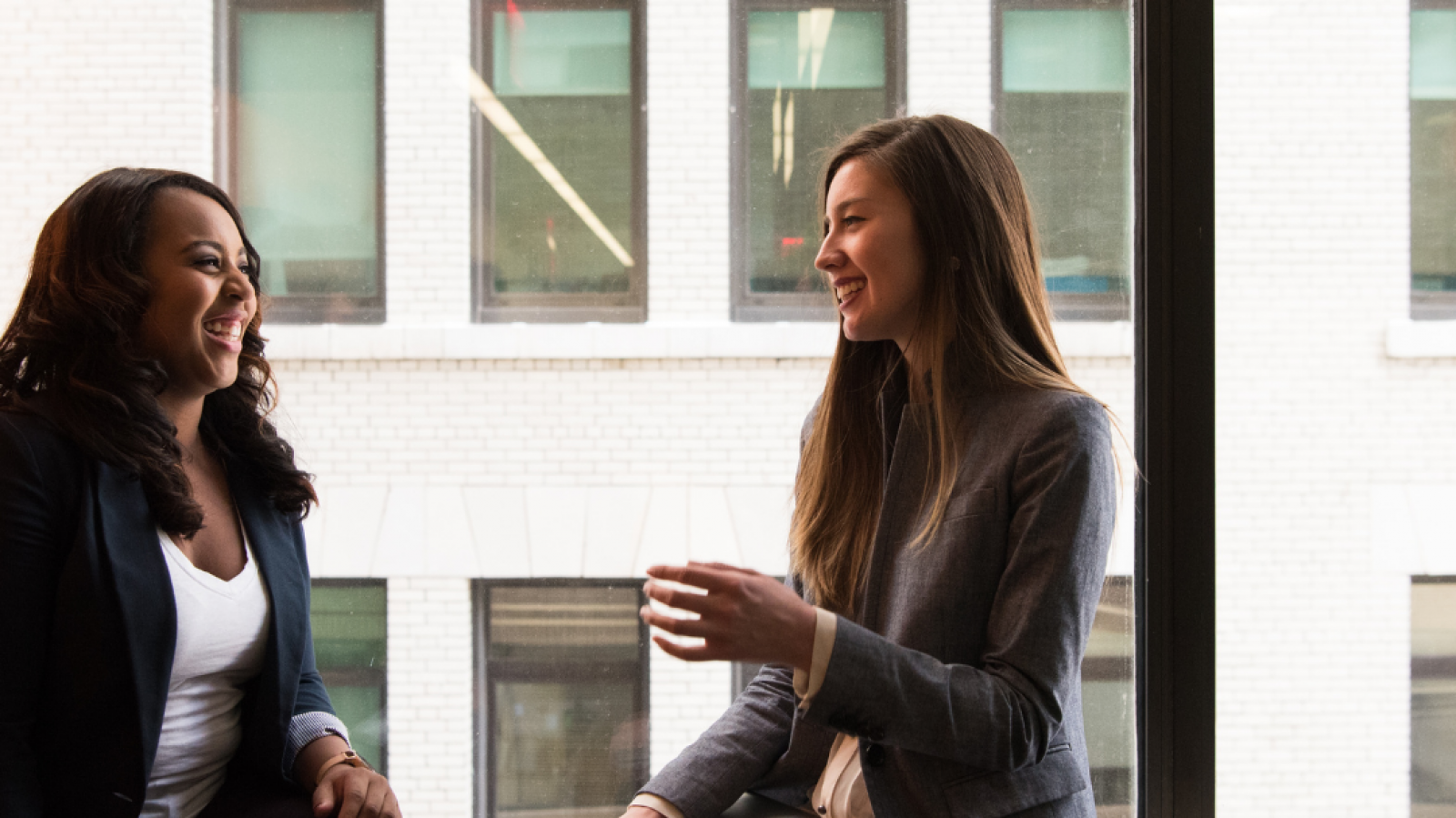 Two ladies speaking to each other by a window in an office setting. 